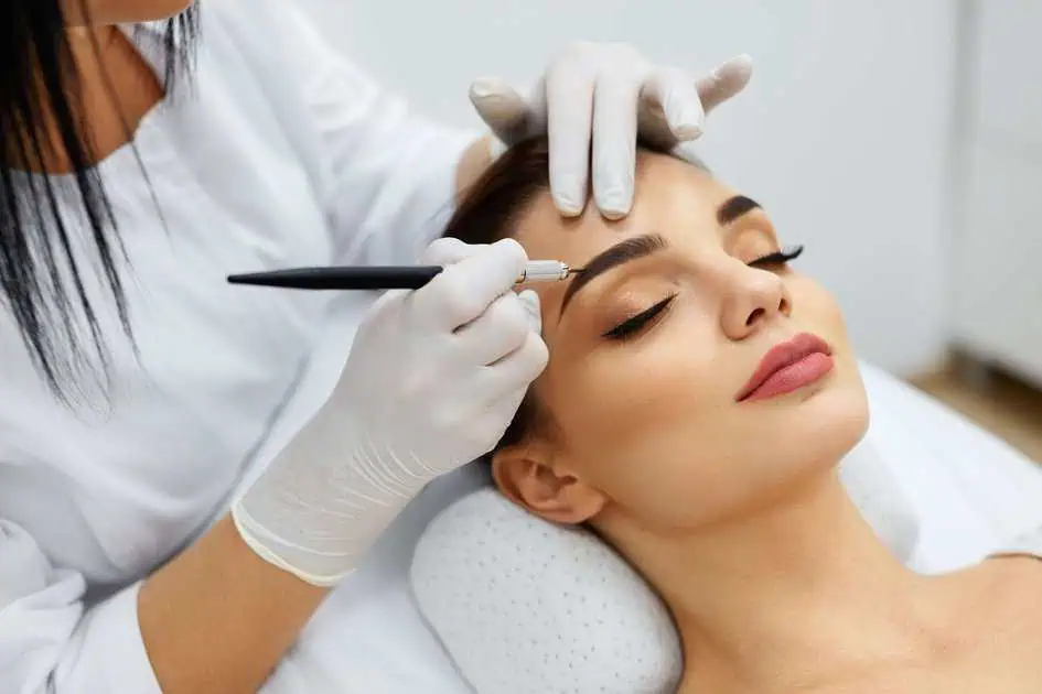 A woman getting eyebrow treatments with a microblading tool.