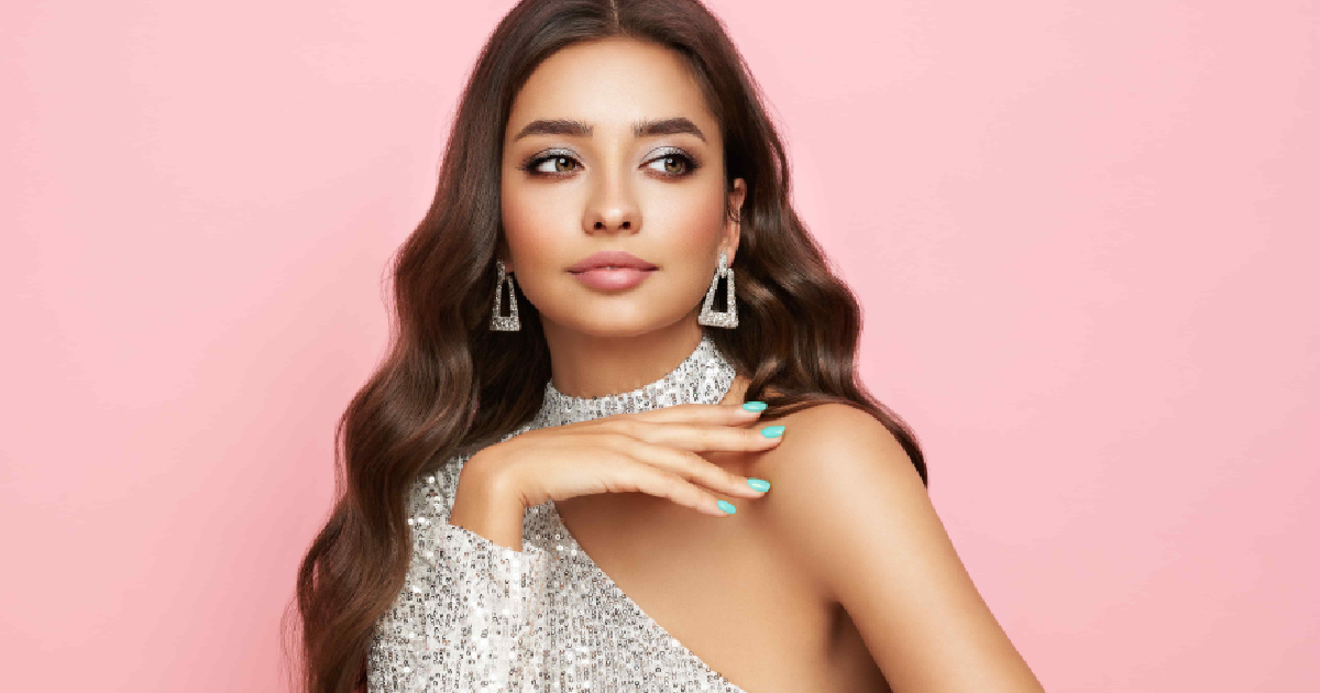 Elegant woman with long wavy brown hair, wearing sparkling silver one-shoulder dress and statement earrings, showcasing permanent jewelry, posing against a pink background in Missoula, MT